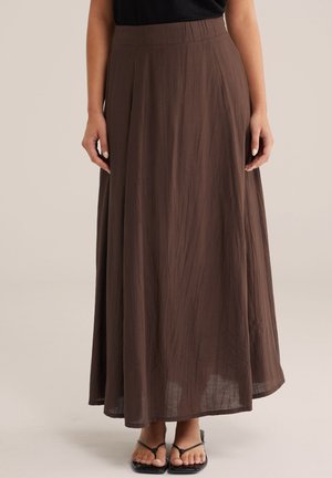 Femeie purtând o fustă maro, până la gleznă, cu textură fluidă, un top negru și sandale negre cu barete, pe un fundal simplu.