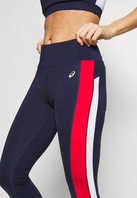 Kvinde justerer navy leggings med røde og hvide lodrette striber på siden og et synligt logo nær taljen.