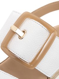 Primer plano de una correa de sandalia de cuero blanco texturizado con una hebilla rectangular translúcida beige y un pasador de metal dorado.