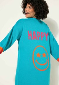 Tyrkysový pletený cardigan s velkým růžovým nápisem "HAPPY" na zadní straně a oranžovou usmívající se tváří se hvězdami. Dlouhé rukávy s oranžovými akcenty.