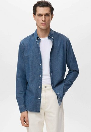 Blue denim shirt with a button-down collar, long sleeves, and white buttons, layered over a white t-shirt and paired with light-colored pants.