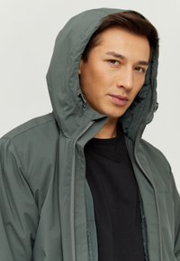 Dunkelgrüne, isolierte Jacke mit Kapuze, ausgestattet mit einem durchgehenden Reißverschluss, elastischen Bündchen und einer glatten, matten Textur. Darunter ein schwarzes T-Shirt.