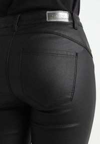 Close-up of black coated jeans with back pocket and silver brand label on waistband against a plain background.