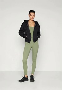 Sudadera negra con cremallera y bolsillos, llevada sobre una camiseta sin mangas ajustada de color verde claro y leggings a juego. Zapatillas deportivas negras completan el conjunto.