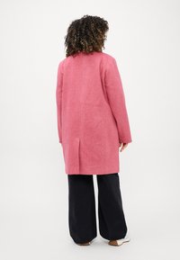 Abrigo de lana rosa con corte recto, abertura en la espalda y mangas amplias, combinado con pantalones negros anchos y zapatillas blancas.