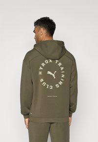 Olivengrønn hettegenser med snorhette, med et sirkulært logo og tekstdesign bak. Mykt stoff med ribbede mansjetter og kant.