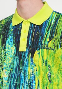 Polo-shirt med en lys gul krave, der har et multifarvet abstrakt mønster i nuancer af blå og grøn, inkluderer frontknapper og broderet logo.