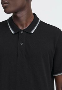 Svart polo skjorte med en tre-knapp placket, hvit og gråstripet krage og korte ermer. Laget av et teksturert stoff.