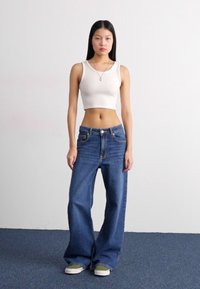 Valge lühike tank top, sinised laiad teksad, rohelised ja valged kanga tossud, seismine sinisel vaibal lihtsa tausta ees.