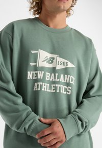 Felpa verde in cotone con maniche lunghe, polsini a coste e un design grafico bianco che presenta "New Balance Athletics" e un motivo a bandiera.