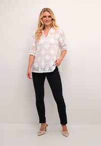 Blouse en dentelle blanche avec motifs floraux, décolleté en V et manches volantes, associée à un pantalon noir ajusté et des mules à talons de couleur claire.