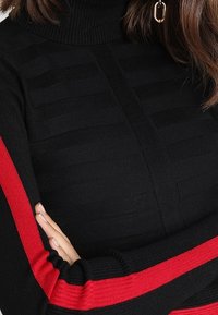 Suéter de cuello alto negro con rayas verticales y detalles rojos a lo largo de las mangas, hecho de un material de punto texturizado.