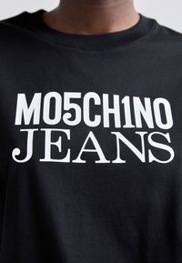 Černé tričko s tučným bílým textem "MO5CH1NO JEANS", které představuje módní prohlášení. Pozadí je jednoduché a neutrální.