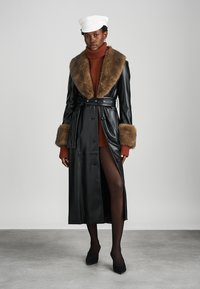 Melnas ādas trenca mētējs ar brūnu mākslīgās kažokādas apkakli un aprocēm; jostas viduklis; saskaņots ar brūnu džemperi un melnām kurpēm.