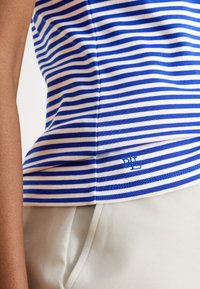 Κοντινό πλάνο μπλε και λευκού ριγέ πουκαμίσου που έχει μπει μέσα σε ανοιχτό μπεζ παντελόνι, με μικρό κεντημένο λογότυπο κοντά στο τελείωμα.