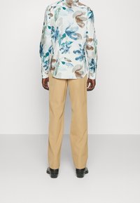 Chemise blanche à manches longues avec des motifs floraux bleus et bruns, associée à un pantalon beige et des chaussures noires, vue de dos.