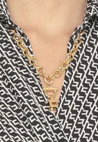 Collana di catena d'oro con un grande pendente a logo, indossata sopra una camicia a scacchi in bianco e nero con scollatura a V.