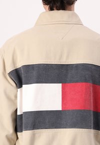 Beige jakke med en fargeblokkert bakdel som har grå, hvite og røde striper. Teksturert stoff med klassisk krage og sømdetaljer.