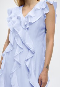 Γυναίκα με αμάνικο φωτεινό γαλάζιο φόρεμα με στρωματοποιημένα βολάν και εμφανή τατουάζ και στους δύο πήχεις.