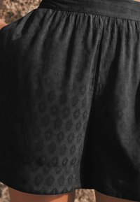 Schwarze Shorts mit einem strukturierten Muster aus Rautenformen, elastischem Bund und lockerer Passform. Das Material wirkt weich und leicht schimmernd.
