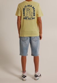 Žluté bavlněné tričko s grafickým potiskem na zadní straně, doplněné světle modrými denimovými šortkami. Model má na sobě černé tenisky s bílými akcenty.