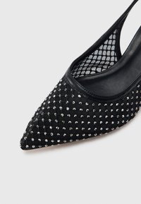 Černá obuv se zadním páskem, s špičatou špičkou, síťovanými boky a ozdobená stříbrnými hroty pro texturu a detaily.