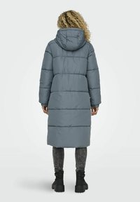 Manteau en duvet gris long avec des surpiqures horizontales, une capuche, et des poches latérales. Porté par-dessus des leggings gris foncé et des bottes noires épaisses.