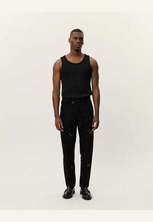Schwarzes, geripptes Tanktop kombiniert mit lässigen, zerrissenen schwarzen Jeans, die mit Patches verziert sind und eine lockere Passform bieten. Getragen mit schwarzen Schnürschuhen.