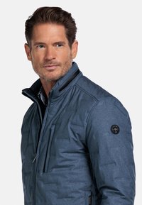 Blauwe, gevoerde jas met een staande kraag, ritssluiting aan de voorkant en een gestructureerd oppervlak. Voorzien van een ritsborstzak en een logo patch op de mouw.