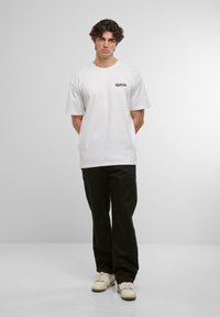 White cotton T-shirt with short sleeves and a crew neckline, featuring black text on the chest. Paired with black pants and off-white sneakers.