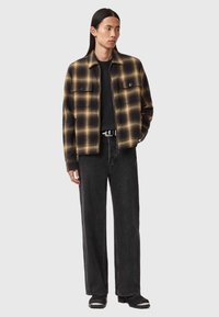 Giacca a quadri nera e marrone con due tasche sul petto, indossata sopra una t-shirt nera e abbinata a jeans neri larghi e stivali neri.