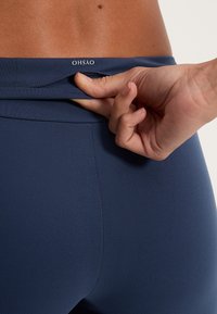 Tengerészeti magas derekú leggings sima textúrával és diszkrét varrási részletekkel. A márka logója a derékpánton látható.