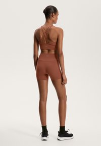 Sport-BH och shorts set i rostfärg, med en korsad ryggdesign, slät textur och minimal varumärkesprofil. Skorna är svarta sneakers.