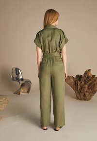 Khaki overal s krátkými ohrnutými rukávy a páskem v pase, s volným střihem a jemnou texturou. Stylizováno s černými sandály.