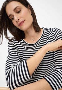 Gestreepte lange mouwen top in marineblauw en wit met een ronde hals. Gladde textuur met contrasterende strepen; model draagt een eenvoudige zilveren ketting.
