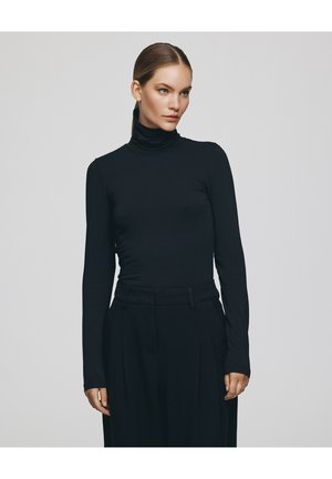 Kobieta o jasnej karnacji i brązowych włosach związanych do tyłu, ubrana w dopasowany czarny golf i czarne spodnie z wysokim stanem na szarym tle.