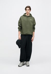 Ελιά πράσινη hoodie με μπροστινή τσέπη και κορδόν�ια, συνδυασμένη με φαρδιά μαύρα παντελόνια και γκρι αθλητικά παπούτσια. Καπέλο κρατημένο στο αριστερό χέρι.