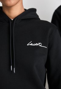 Asmuo vilkintis juodą kapučiną su balta Lacoste logotipu ant krūtinės ir raišteliais su Lacoste ženklu.