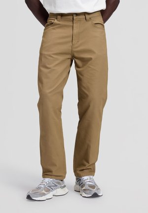 Mann trägt hellbraune gerade geschnittene Hose und graue New Balance-Sneakers, steht vor einem schlichten hellgrauen Hintergrund.