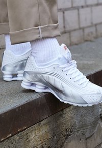 Baskets Nike blanches et argentées portées avec des chaussettes blanches et un pantalon beige, posant le pied sur le bord d'un rebord en béton à l'extérieur.