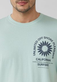  Lichtblauwe katoenen t-shirt met korte mouwen en ronde hals; heeft marineblauwe tekst en grafisch ontwerp met een stralend patroon aan de voorkant.