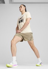Lyse beige t-shirt med trykt design, parret med khaki shorts med sidelommer. Hvide sko med gule og lyserøde accenter.