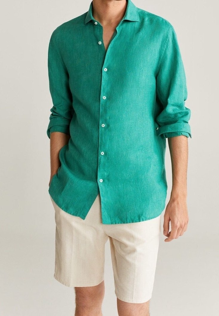 Homme portant une chemise bleu sarcelle à manches longues avec des poignets retroussés et un short beige jusqu'aux genoux sur un fond uni.