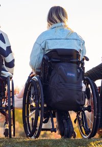 Des fauteuils roulants avec des roues noires, équipés d'un sac à dos gris, placés sur de l'herbe. Une personne porte une veste en jean et des chaussures blanches.