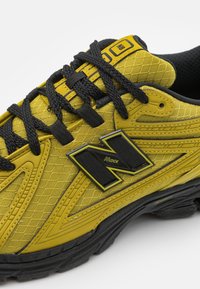 Žlutá sportovní obuv s černými doplňky, vzorovaným svrchem, texturovaným síťovinovým materiálem a výrazným logem "N" na boku. Součástí jsou černé tkaničky.