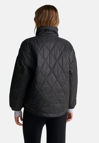 Černá prošívaná bunda s vysokým límcem, která má diamantový vzor, volný střih a žebrované manžety. Měkká, lesklá tkanina s minimální texturou.