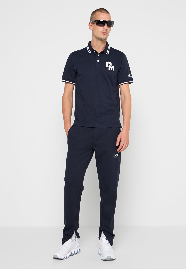 Granatowa koszulka polo z kontrastującymi paskami, z logo na piersi; zestawiona z dopasowanymi dresami. Obie wykonane z miękkiego, elastycznego materiału.