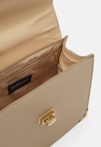 Beige syntetisk håndtaske med en glat tekstur, en guld metalspænde og en indvendig lynlås lomme med stofforing. Brandmærket synligt.