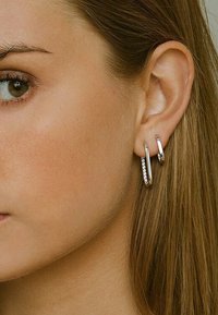 Orecchini a cerchio in argento con una finitura liscia e una fila di piccole pietre trasparenti su uno di essi. Design leggero, indossati all'orecchio di una donna.