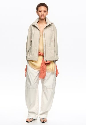 Persona con chaqueta beige sobre una blusa amarilla con ribete de encaje, camisa naranja atada a la cintura, pantalones blancos sueltos y chanclas negras, sobre fondo blanco.